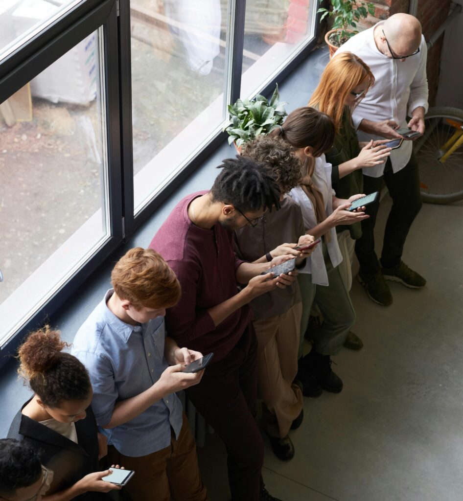 A group of people standing by a window, each using their smartphones, representing the connected world and the benefits of social media marketing in engaging audiences.